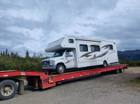 A semi-truck from Help Line Towing transporting an RV on a flatbed trailer under a cloudy sky in Wasilla, AK.