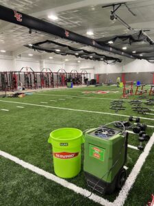 Servpro drying equipment, including a dehumidifier and bucket, deployed on an indoor sports field for water damage restoration in Athens, AL.