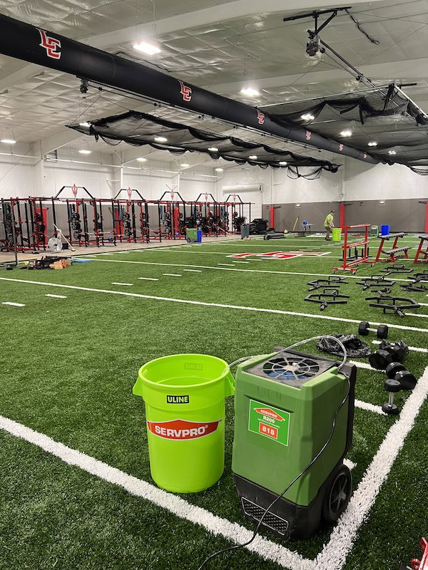 Servpro drying equipment, including a dehumidifier and bucket, deployed on an indoor sports field for water damage restoration in Athens, AL.