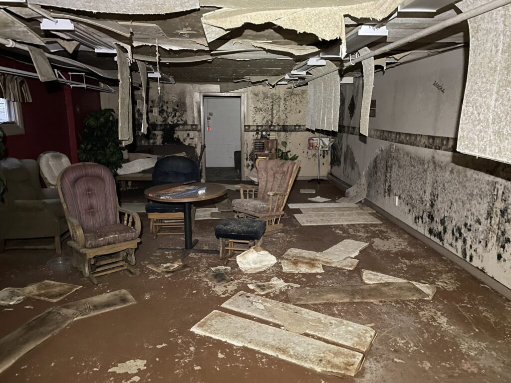 A severely water-damaged basement with collapsed ceiling tiles, mold growth on walls, and debris on the floor, requiring restoration by The Restoration Experts in Waupaca, WI.