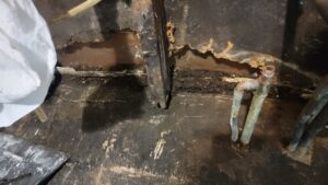 A close-up of severe water damage on a floor, showing dark, possibly moldy wood and exposed pipes, a restoration project for Ciocca Cleaning & Restoration in Fort Wayne, IN.
