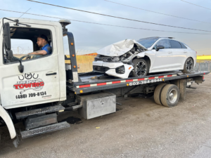 A severely damaged white car being transported on a flatbed tow truck by Trejo's Towing LLC in Phoenix, AZ.
