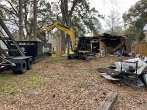 An excavator from Judice Dirt & Demolition using a grapple to load debris from a demolished shed into a dumpster in Lafayette, LA.