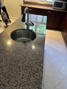 A shiny kitchen island with a clean sink and granite countertop, with cleaning equipment visible, by Simply Clean Jax in Jacksonville, FL.