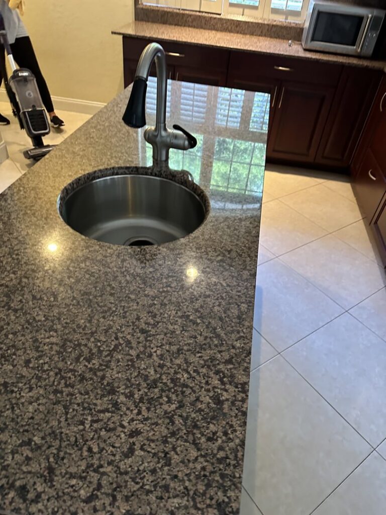 A shiny kitchen island with a clean sink and granite countertop, with cleaning equipment visible, by Simply Clean Jax in Jacksonville, FL.