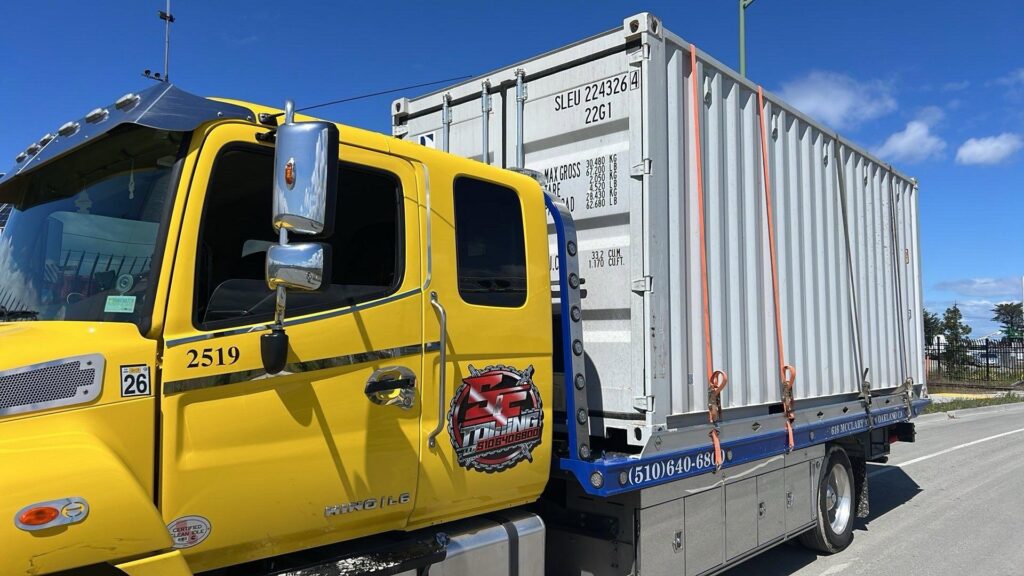 A large shipping container is being transported on a flatbed tow truck by S&F Towing in Oakland, CA.