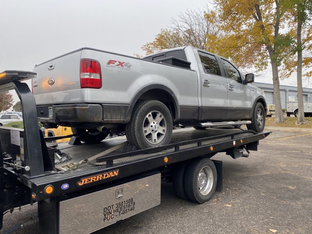 A silver Ford F-150 pickup truck being transported on a flatbed tow truck by Black Mamba Towing & Recovery Llc in Aurora, CO.