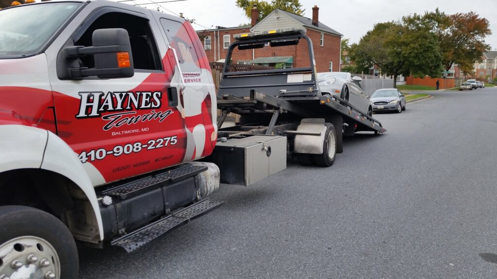 A silver Hyundai Veloster being transported on a flatbed tow truck at night by Baltimore Towing Service LLC in Owings Mills, MD.