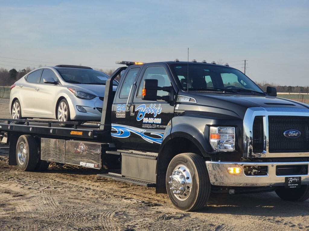 A silver sedan loaded and secured on a Bobbys Towing & Recovery flatbed tow truck in Raleigh, NC.