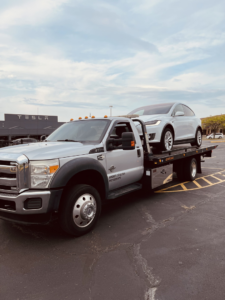 A silver flatbed tow truck transporting a white Tesla Model X for FE Transportation LLC in Cincinnati, OH.