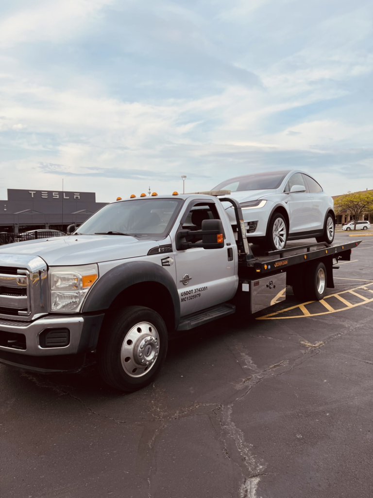 A silver flatbed tow truck transporting a white Tesla Model X for FE Transportation LLC in Cincinnati, OH.