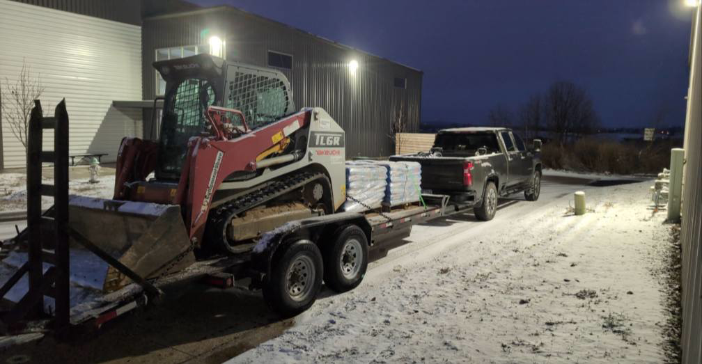 A skid steer loaded on a trailer, towed by a truck on a snowy night for Creative Grounds in Springdale, AR