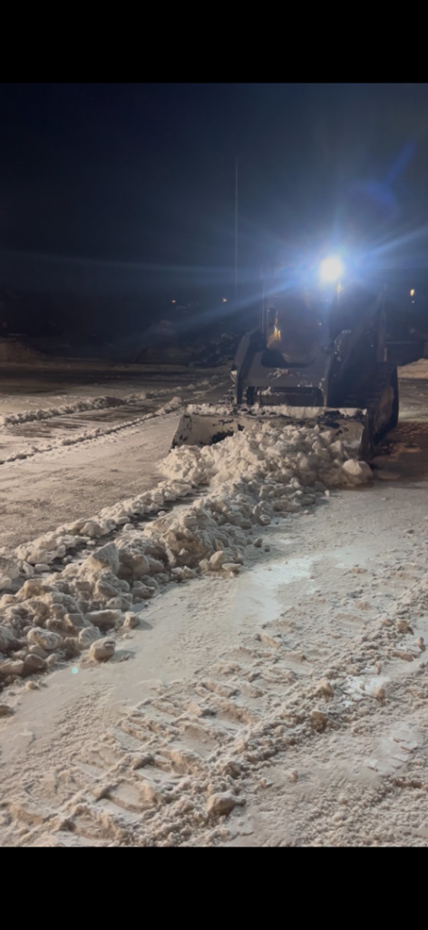 A skid steer with a plow attachment actively clearing snow from a parking lot at night by OnPoint Land Solutions, LLC in Enid, OK.