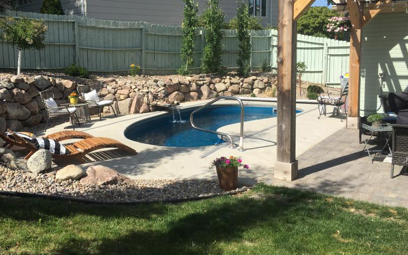 A small kidney-shaped swimming pool with a waterfall feature and surrounding patio by Backyard Escapes by Kerner Pools & Spas in Bismarck, ND.