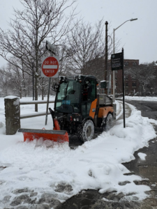 A small plow vehicle clearing a snowy path in a park for the City of Cambridge Department of Public Works in Cambridge, MA.