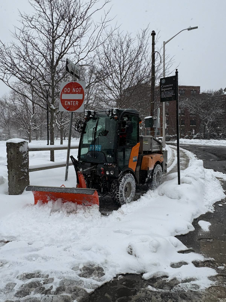 A small plow vehicle clearing a snowy path in a park for the City of Cambridge Department of Public Works in Cambridge, MA.