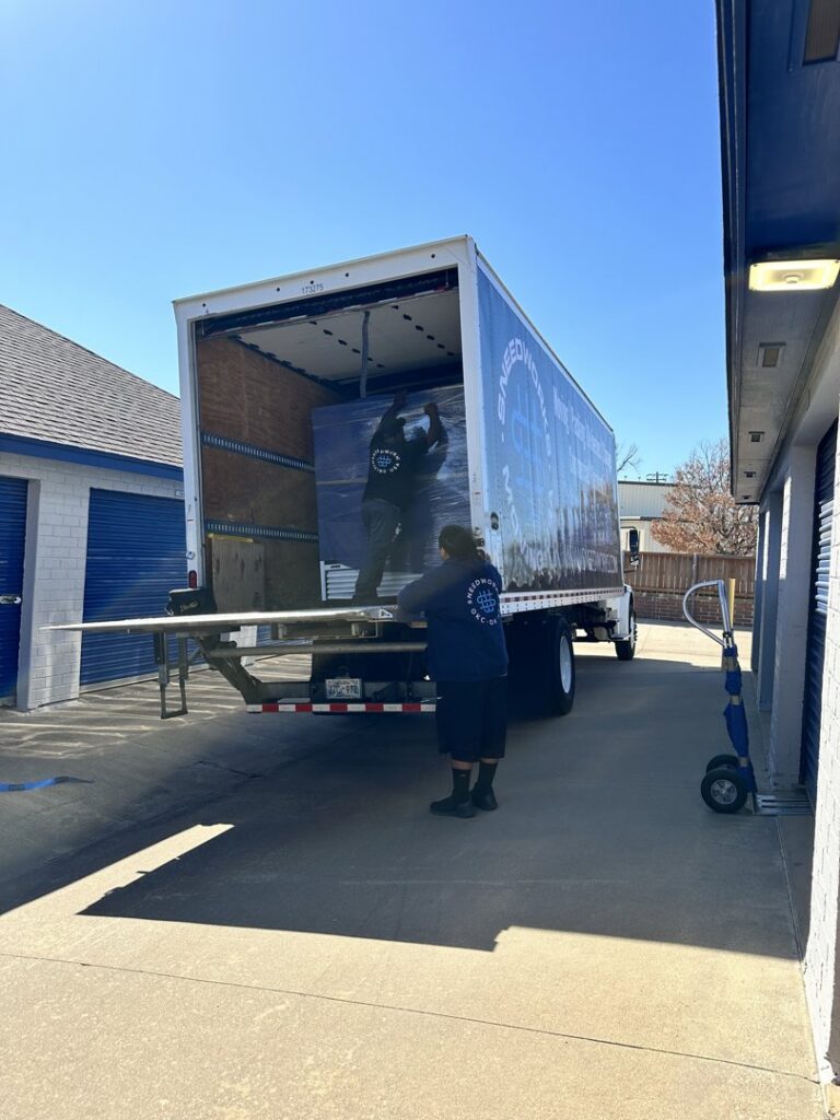 Two SNEEDWORKS movers loading a large, wrapped item onto a moving truck near storage units in Oklahoma City, OK.