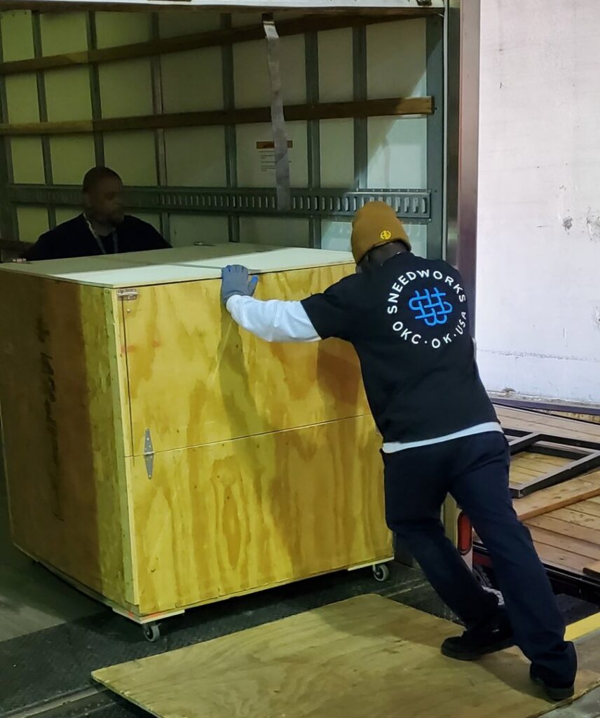 Two SNEEDWORKS movers pushing a large wooden crate onto a moving truck in Oklahoma City, OK.