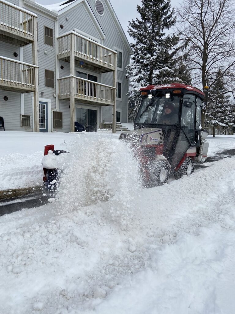 A compact utility tractor with a snow blower attachment actively clearing snow from a residential driveway for C&S Lawncare in Ann Arbor, MI.