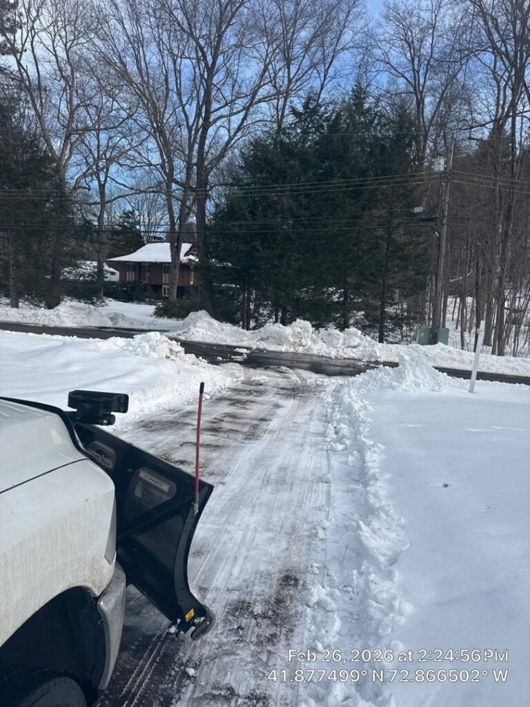 A white pickup truck with a snow plow clearing a residential driveway, with snow piled high, by Diamond Image in New Haven, CT.