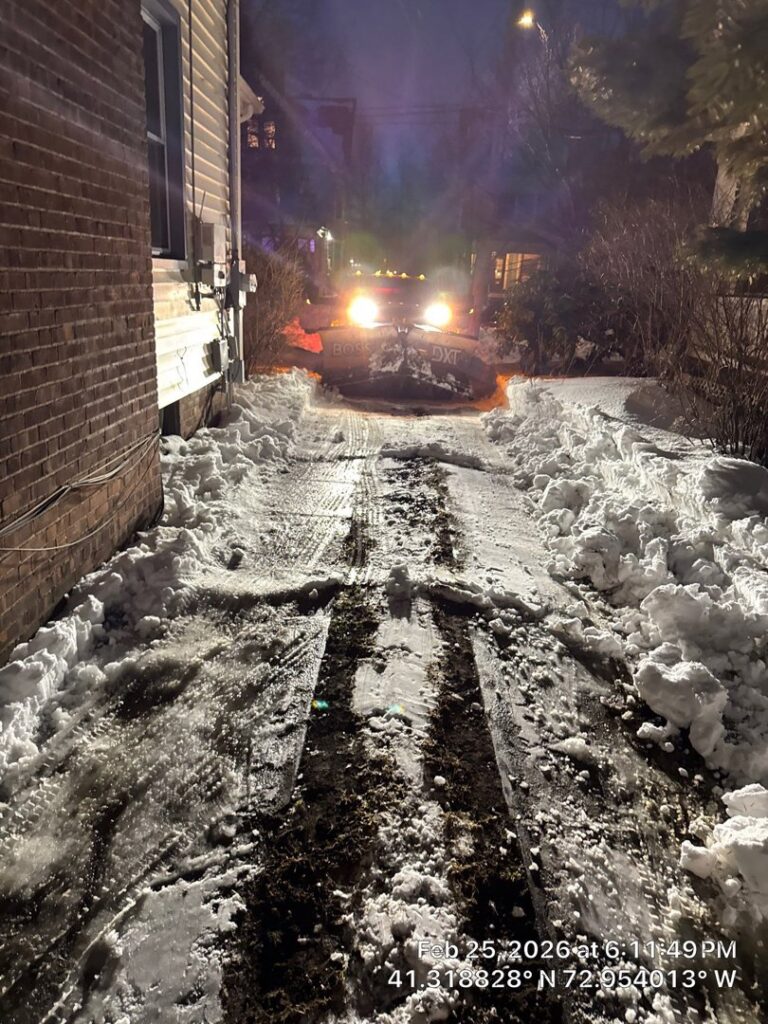 A white pickup truck with a snow plow clearing a snowy residential driveway at night, by Diamond Image in New Haven, CT.