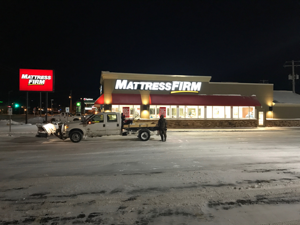 A snow plow truck with a salt spreader working in a commercial parking lot at night for Appleton Snow Removal in Appleton, WI.
