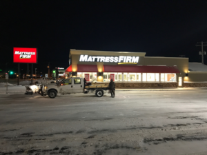 A snow plow truck with a salt spreader working in a commercial parking lot at night for Appleton Snow Removal in Appleton, WI.