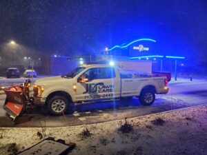 A JD Lawn Care LLC snow plow truck clearing snow from a commercial parking lot at night in Berea, OH.
