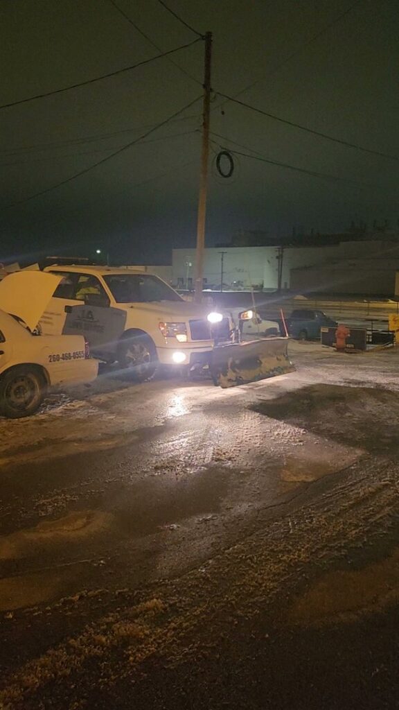 A JA Lawn Service, LLC truck with a snow plow actively clearing snow from an area at night in Fort Wayne, IN.