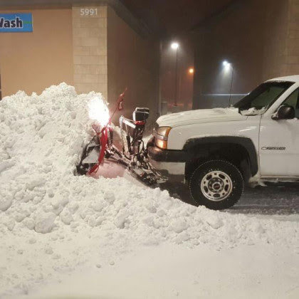 A white pickup truck with a red snow plow actively clearing a large pile of heavy snow at night, performed by LAWN Sharks LLC in Colorado Springs, CO.