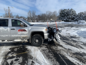 A snow plow truck from American Snow Removal LLC clearing a commercial parking lot in Des Moines, IA, with piles of snow visible.