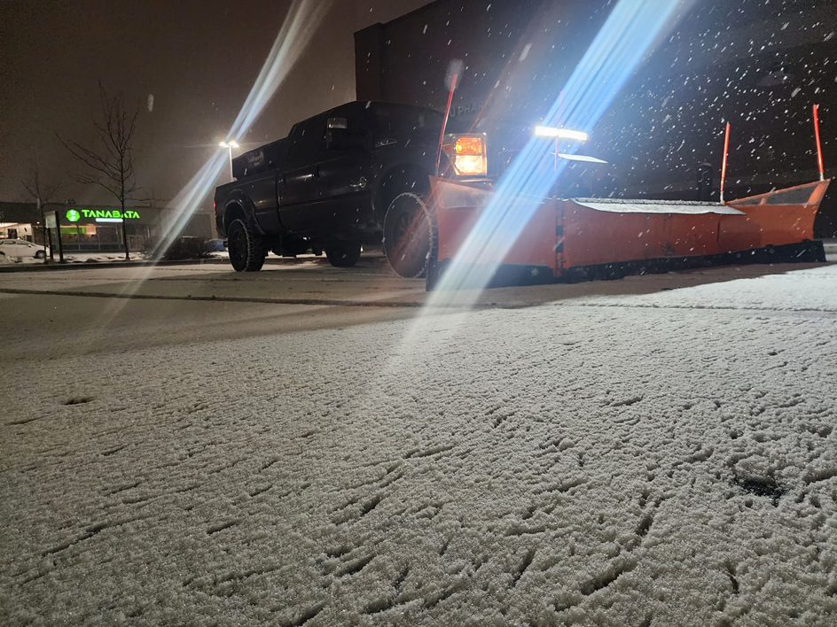 A snow plow truck with its lights on, clearing a parking lot at night during a snowfall for Mr Plow in South Salt Lake, UT.