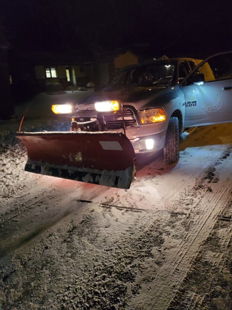Snow plow truck with headlights on, clearing snow from a residential street at night by Brinkley'lawn service in Brooklyn Park, MN.