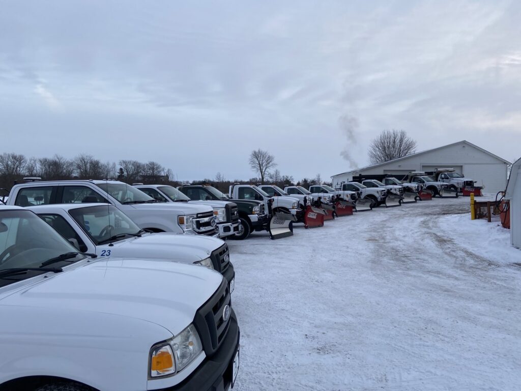 A line of pickup trucks equipped with snow plows, parked in a snowy lot, ready for snow removal by C&S Lawncare in Ann Arbor, MI.
