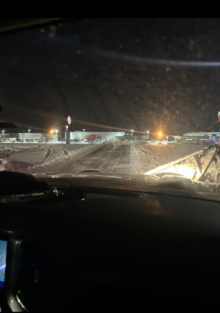 A snow plow clearing a commercial parking lot at night, provided by First Class Lawn Care and Snow Removal LLC in Shawnee, KS.