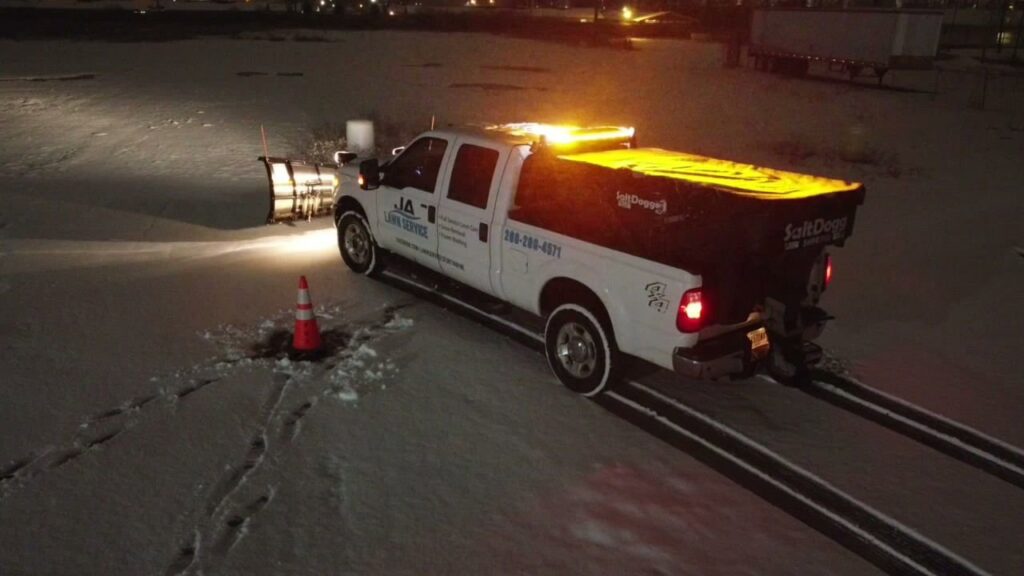 A JA Lawn Service, LLC truck with a snow plow and salt spreader clearing a snowy parking lot at night in Fort Wayne, IN.