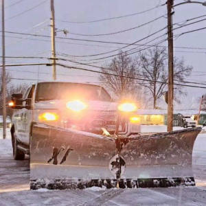 A JA Lawn Service, LLC truck actively plowing snow from a road in Fort Wayne, IN, with its headlights on.
