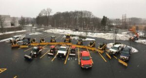 A fleet of snow removal trucks and skid steers with plows ready for work in a snowy parking lot by The Groundskeeper Inc. in Fort Collins, CO.