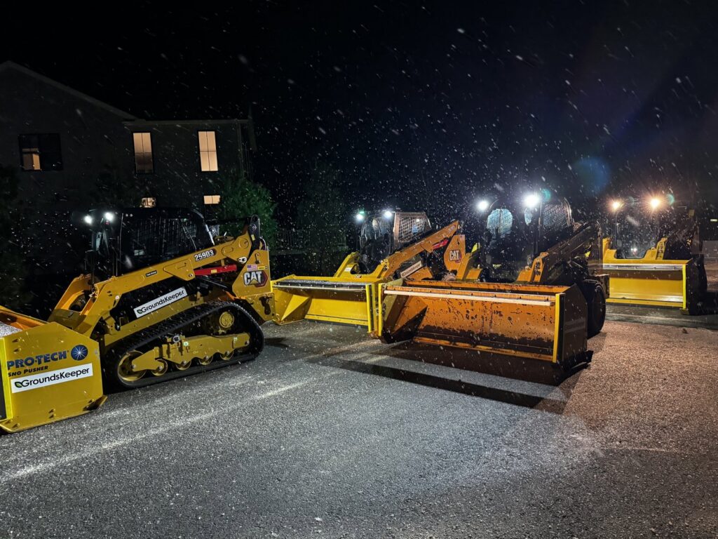 Skid steers with snow pushers lined up at night during snowfall, ready for snow removal by The Groundskeeper Inc. in Fort Collins, CO.