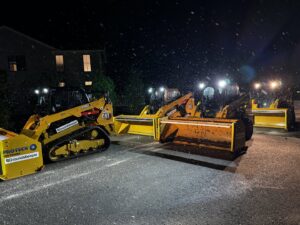 Skid steers with snow pushers lined up at night during snowfall, ready for snow removal by The Groundskeeper Inc. in Fort Collins, CO.