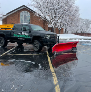 A snow removal truck with a red plow and a spreader clearing a parking lot for Earthscapes Luxury Curb & Landscape in Layton, UT.