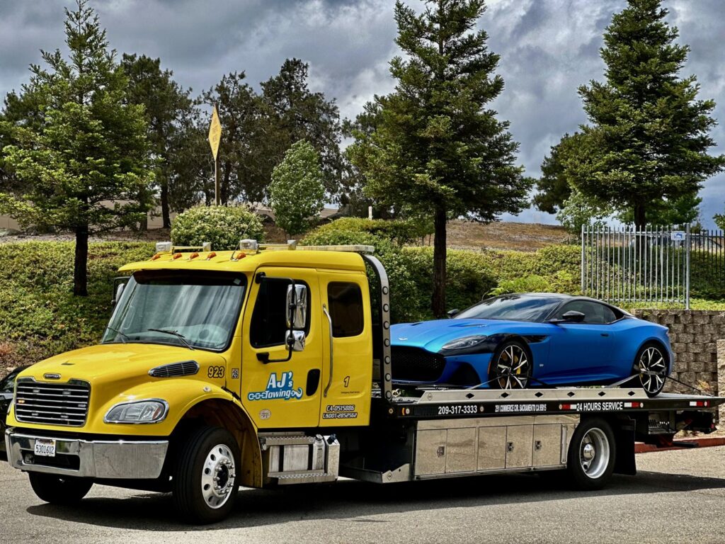 A yellow flatbed tow truck from AJ's Towing Services & Roadside Assistance towing a blue sports car in Sacramento, CA.
