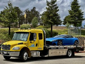 A yellow flatbed tow truck from AJ's Towing Services & Roadside Assistance towing a blue sports car in Sacramento, CA.