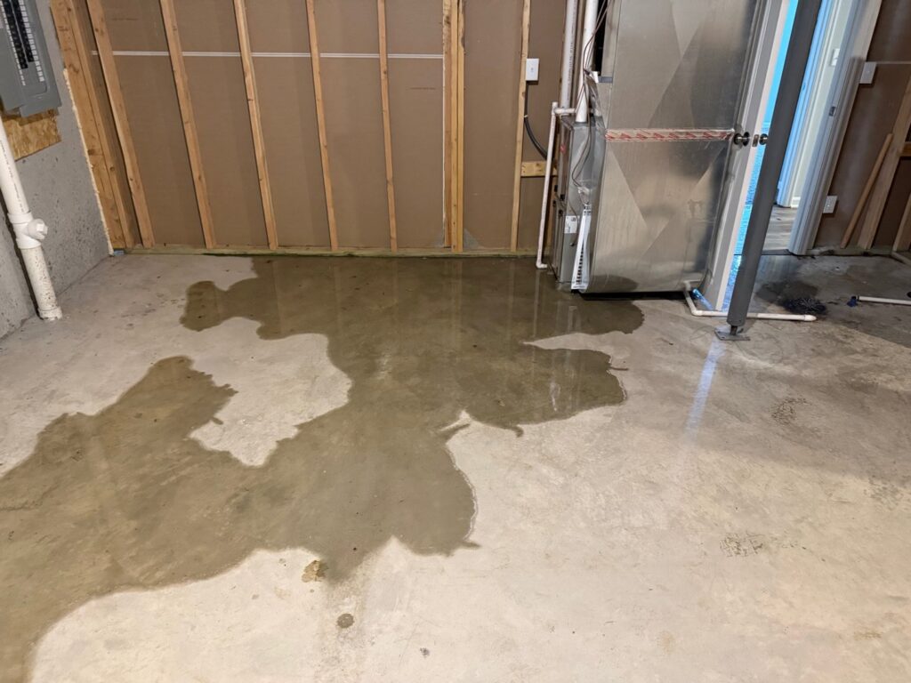 Standing water on a concrete basement floor next to a furnace, indicating water damage, addressed by Cowboy Certified Plumbing & Water Cleanup in Lee's Summit, MO.