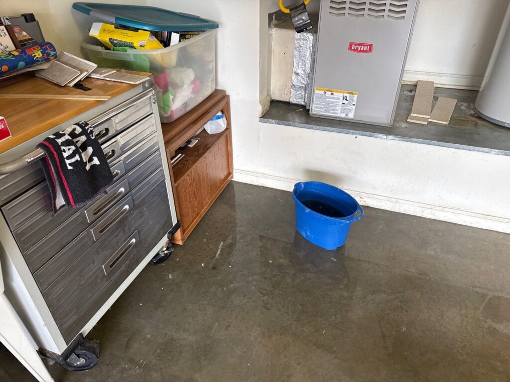 Standing water on a garage or utility room floor with a blue bucket, indicating active water damage, by PuroClean of Hilton Head in Bluffton, SC.