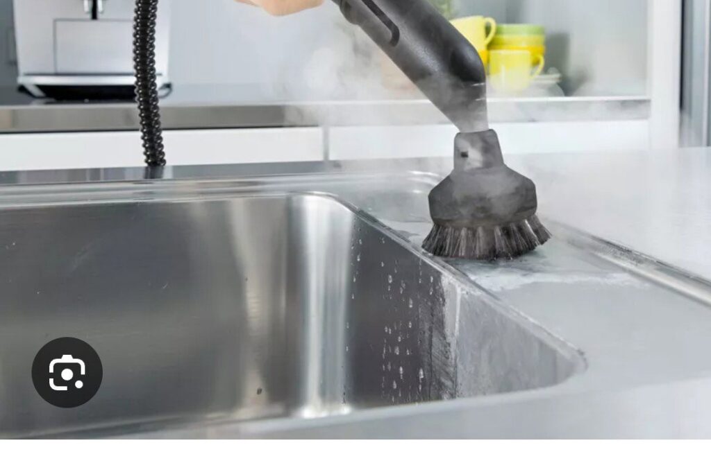 A hand using a steam cleaner with a brush attachment to clean a stainless steel kitchen sink by D A E S Cleaning LLC in Billings, MT.
