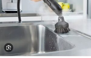 A hand using a steam cleaner with a brush attachment to clean a stainless steel kitchen sink by D A E S Cleaning LLC in Billings, MT.