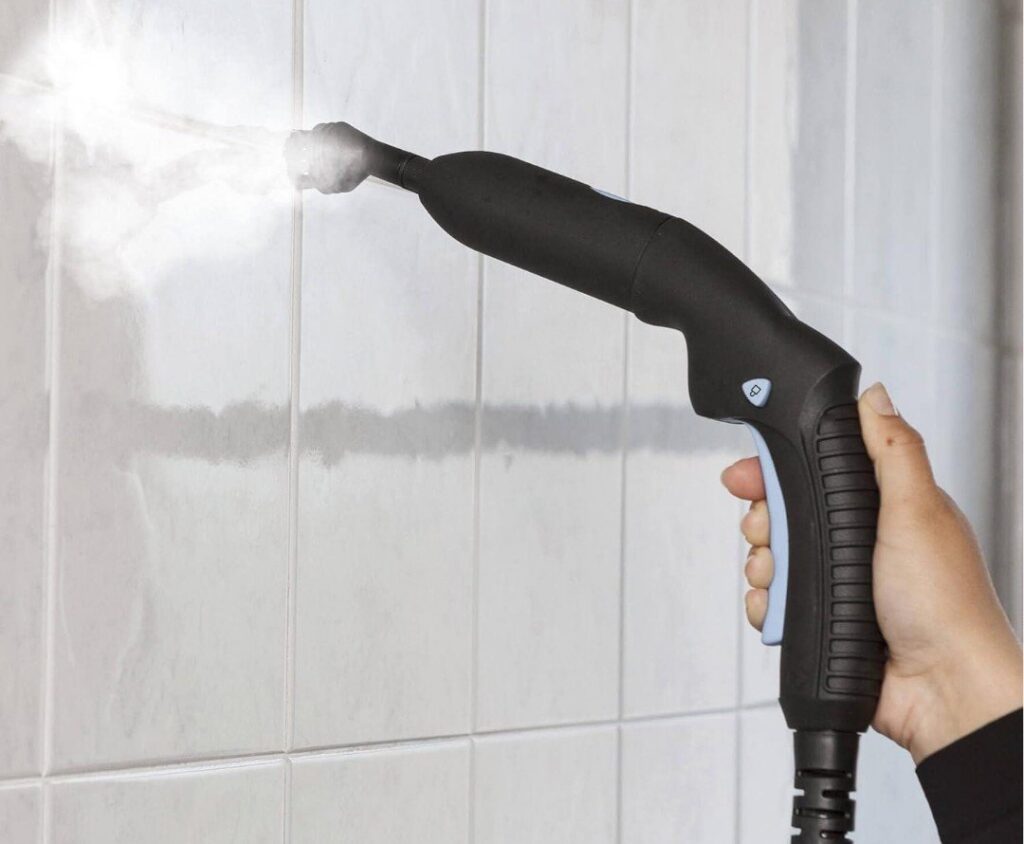 A hand using a steam cleaner to clean a white tiled wall, likely in a bathroom or kitchen, by D A E S Cleaning LLC in Billings, MT.