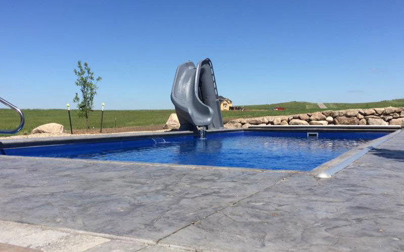 A swimming pool featuring a slide and stamped concrete decking by Backyard Escapes by Kerner Pools & Spas in Bismarck, ND.