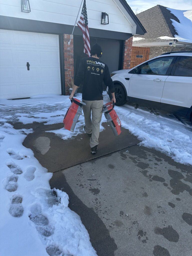 A Paul Davis technician carrying drying equipment through a snowy driveway, ready for a water damage job in Norman, OK.